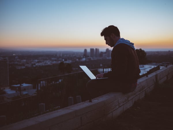 A man using his laptop on a ledge at sunset.